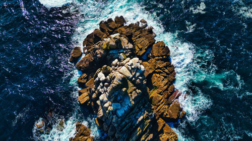 Aerial view of a rocky coastline with waves crashing against the rocks. The water is deep blue with white foam from the waves, highlighting the rugged texture of the rocks.