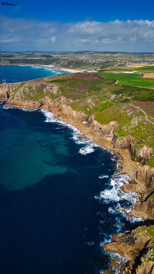 Aerial view of a rugged coastline with cliffs, clear blue waters, and a sandy beach in the distance. The landscape features green fields and a small village, under a bright blue sky with scattered clouds.