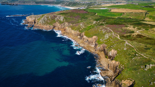 Aerial view of a rugged coastline with cliffs, clear blue waters, and green fields. The landscape features rocky outcrops and a winding path along the cliffs, with a distant view of a coastal village.