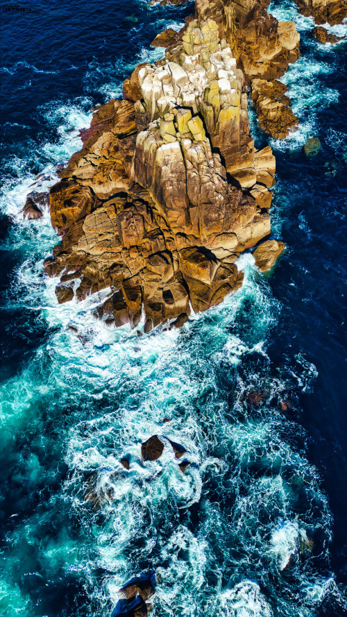 Aerial view of rugged coastal rocks surrounded by turquoise ocean waves. The water crashes against the rocky formations, creating white foam. The scene captures the beauty of nature with a vibrant contrast between the rocks and the sea.
