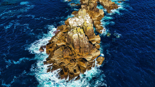 Aerial view of rocky coastline with waves crashing against the rocks. The water is deep blue, contrasting with the rugged, textured rocks. The scene captures the beauty of nature and the power of the ocean.