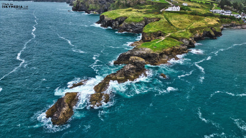 Aerial view of a rocky coastline with lush green hills and white houses on the cliffs. Waves crash against the rocks, creating foam in the turquoise water. The landscape is scenic and tranquil, showcasing the beauty of nature.