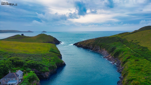A scenic coastal view featuring a narrow waterway surrounded by lush green hills. The sky is partly cloudy with hints of blue peeking through. In the foreground, a charming house is visible near the water's edge, while distant islands can be seen on the horizon.