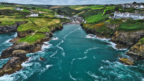 A scenic coastal view featuring rocky cliffs, lush green hills, and a tranquil blue sea. Small white houses dot the landscape, nestled among the greenery, with waves gently crashing against the shore.