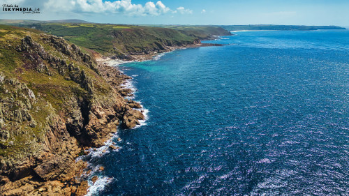 Aerial view of a rugged coastline with cliffs and clear blue waters. The landscape features green hills in the background, rocky shores, and a serene ocean. The sky is partly cloudy, enhancing the natural beauty of the scene.