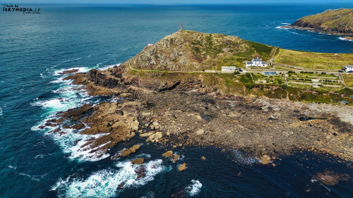 Aerial view of a coastal landscape featuring rocky shores, a lighthouse on a hill, and waves crashing against the rocks. The scene captures the beauty of nature with a clear blue sky and vibrant green hills.
