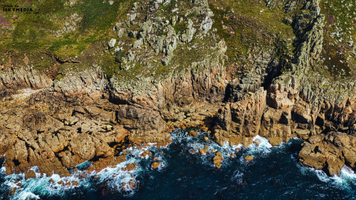 Aerial view of rugged coastline featuring rocky cliffs and crashing waves. The landscape showcases a mix of green vegetation on the cliffs and deep blue ocean waters, creating a dramatic natural scene.