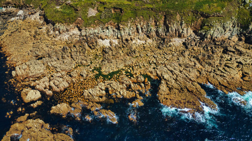 Aerial view of rocky coastline with clear blue water crashing against the shore. The landscape features rugged cliffs and scattered boulders, showcasing a mix of textures and colors from the rocks and vegetation.