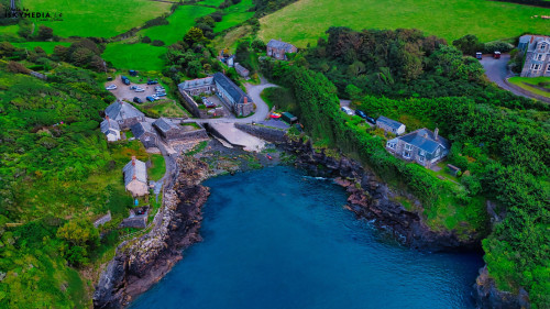 Aerial view of a coastal village with lush green hills, rocky shoreline, and a small harbor. The scene includes several buildings, a parking area, and boats in the water, surrounded by vibrant greenery.