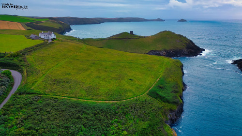 A scenic coastal landscape featuring lush green fields, a winding path, and a serene blue ocean. The coastline is rugged with cliffs and small hills, and a distant tower can be seen on the hilltop. The sky is partly cloudy, adding to the tranquil atmosphere.