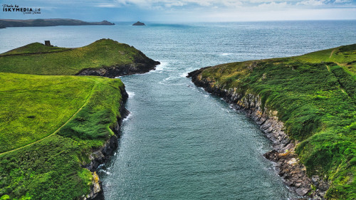 Aerial view of a serene coastal landscape featuring green hills, a narrow waterway, and the ocean in the background. The scene captures the beauty of nature with rocky cliffs and lush vegetation.