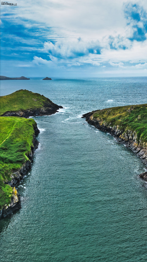 A scenic view of a coastal landscape featuring two green hills flanking a narrow waterway leading into the ocean. The sky is partly cloudy, and the water is calm, reflecting the surrounding greenery.