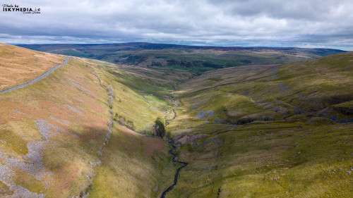 Aerial view of a vast, undulating landscape with green hills, a winding road, and overcast skies.