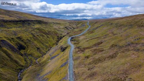 Aerial view of a vast, undulating landscape with green hills, a winding road, and overcast skies.