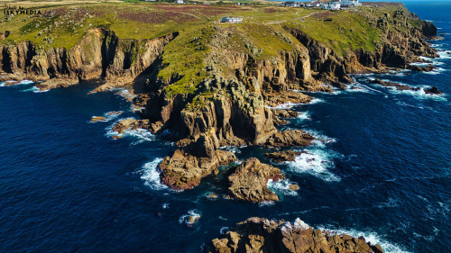 Aerial view of rugged cliffs and rocky coastline with waves crashing against the shore. Lush green hills are visible above the cliffs, and a small white building is perched on the edge. The deep blue ocean contrasts with the rocky landscape.