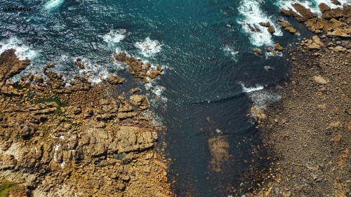 Aerial view of rocky coastline with waves crashing against the shore. The contrast between the dark water and light-colored rocks creates a striking visual. The scene captures the natural beauty of the ocean and rugged terrain.