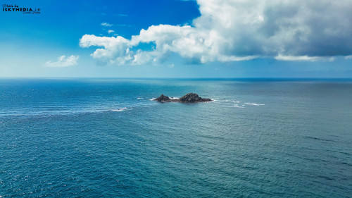 A serene view of a small rocky island surrounded by calm blue ocean waters under a bright sky with fluffy clouds. The horizon stretches far into the distance, creating a peaceful and tranquil atmosphere.