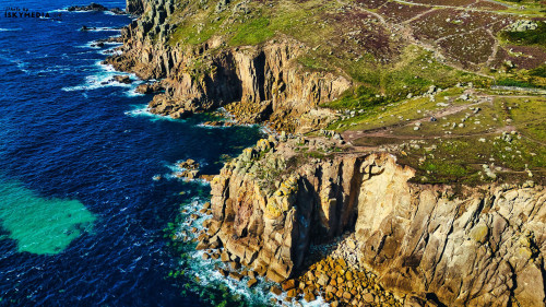 Aerial view of rugged coastal cliffs meeting the ocean, with vibrant blue waters and rocky shoreline. The landscape features green vegetation on the cliffs and a clear sky, showcasing the natural beauty of the coastline.