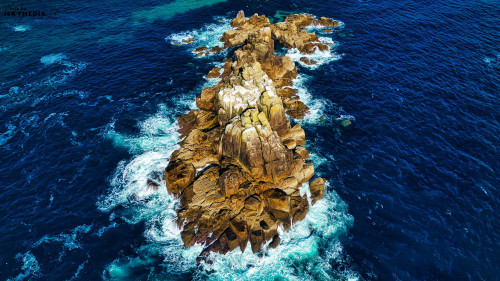 Aerial view of a rocky island surrounded by deep blue ocean waters. Waves crash against the rocks, creating white foam. The island features rugged terrain with patches of greenery on top.