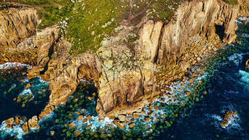 Aerial view of rugged coastal cliffs with green vegetation and rocky shoreline. The ocean waves crash against the rocks, creating a dynamic seascape. Clear blue water contrasts with the earthy tones of the cliffs and rocks.