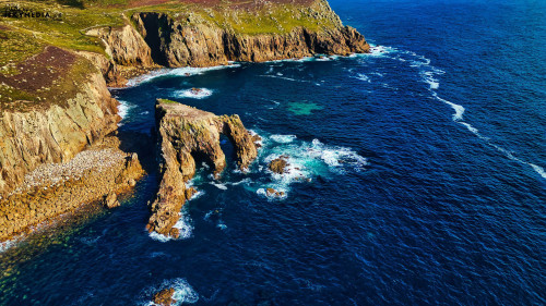 Aerial view of rugged coastal cliffs with a rock formation jutting into the ocean. The water is deep blue with white frothy waves crashing against the rocks. Lush green vegetation covers the cliffs, contrasting with the rocky shoreline.