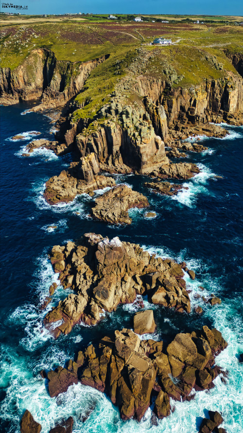 Aerial view of rugged coastal cliffs with rocky formations and crashing waves. The landscape features green hills and a clear blue ocean, showcasing the natural beauty of the coastline.