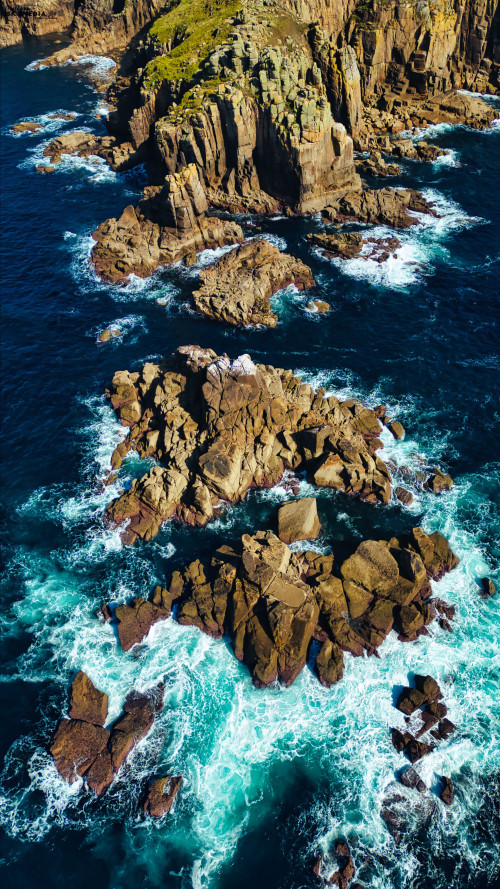 Aerial view of rocky coastline with waves crashing against the rocks. The scene features a mix of dark blue ocean water and rugged, textured rock formations, creating a dramatic natural landscape.