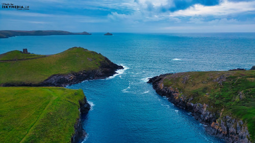 Aerial view of a serene coastal landscape featuring green hills, rocky shores, and a calm blue sea. The horizon is dotted with small islands under a cloudy sky, creating a tranquil and picturesque scene.