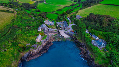 Aerial view of a coastal village surrounded by lush green fields and rocky shores. The village features quaint buildings, a small harbor, and a winding road leading to the water. The landscape is vibrant with greenery and the ocean is calm.