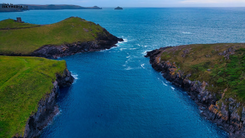 Aerial view of a serene coastal landscape featuring lush green hills and a calm blue sea. The water flows between two rocky formations, with distant islands visible on the horizon. The scene captures the beauty of nature and tranquility.