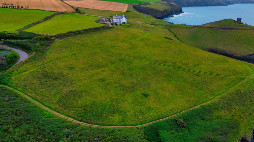 Aerial view of a green landscape with a solitary white house near the coast. The area features rolling hills, agricultural fields, and a winding road leading to the house. The ocean is visible in the background, creating a serene coastal scene.