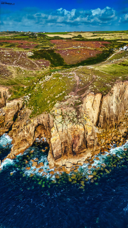Aerial view of rugged cliffs meeting the ocean, with lush green hills in the background. The coastline features rocky formations and clear blue waters, under a bright sky with scattered clouds.