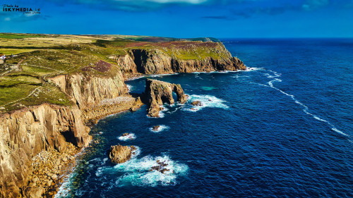 Aerial view of rugged cliffs along a coastline with waves crashing against the rocks. The landscape features green hills and a clear blue sky, showcasing the natural beauty of the seaside.