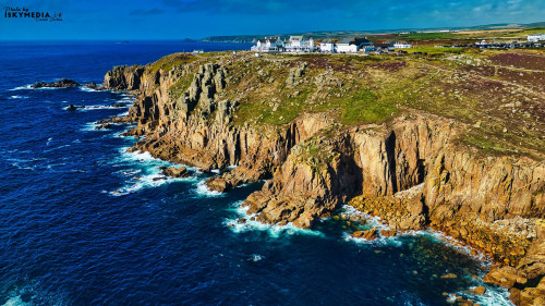A stunning coastal view featuring rugged cliffs meeting the deep blue ocean. The landscape is dotted with green vegetation and a few white buildings perched on the cliffs, under a clear blue sky. Waves crash against the rocky shore, creating a picturesque scene.