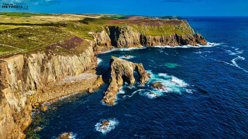 Aerial view of rugged cliffs meeting the ocean, with waves crashing against the rocks. The landscape features green hills and a clear blue sky, showcasing the natural beauty of the coastline.