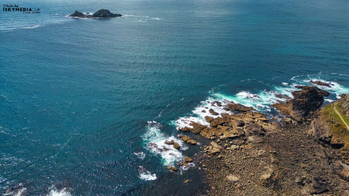 Aerial view of a rocky coastline with clear blue waters and gentle waves. An isolated rocky island is visible in the distance, surrounded by the ocean. The shoreline features a mix of rocks and sandy areas, creating a natural landscape.