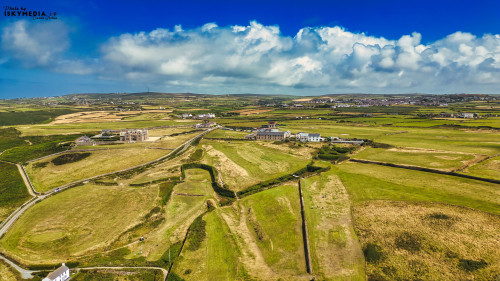 Aerial view of a lush green landscape with rolling hills, fields, and a few scattered buildings under a bright blue sky with fluffy white clouds. The scene captures the beauty of rural countryside, showcasing the vibrant colors of nature.