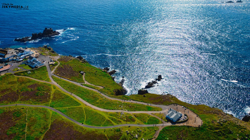 Aerial view of a coastal landscape featuring green hills, winding paths, and a sparkling blue ocean. Small buildings are visible near the shore, with rocky outcrops in the water. The scene captures the beauty of nature and coastal living.