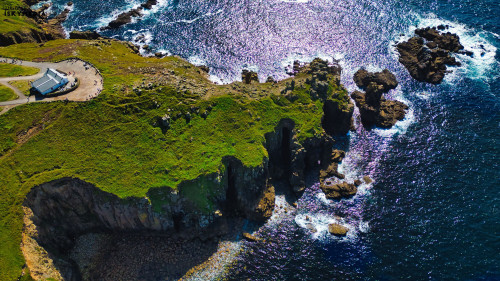 Aerial view of a rugged coastline with cliffs and rocky formations. The ocean is shimmering under sunlight, and a small building is visible on the grassy land. The scene captures the beauty of nature and the coastline's dramatic landscape.