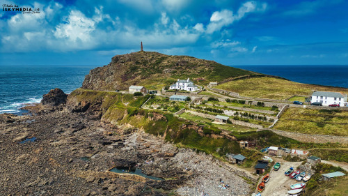 Aerial view of a coastal landscape featuring a rocky shore, a lighthouse on a hill, and a cluster of white buildings. The scene includes boats on the beach and lush greenery, with dramatic clouds in the blue sky above.