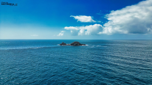 A serene view of a small rocky island surrounded by calm blue ocean waters under a clear sky with fluffy white clouds. The sunlight reflects off the water, creating a tranquil atmosphere.