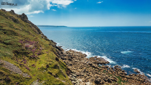 A scenic coastal view featuring rocky cliffs and a vibrant blue ocean under a clear sky. The foreground shows rugged terrain with patches of green and purple vegetation, while the horizon stretches into the distance with a few distant landforms.