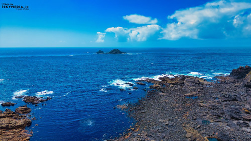 A stunning coastal view featuring a rocky shoreline and clear blue ocean waters. The horizon showcases a few small islands and a bright blue sky with fluffy white clouds. The scene captures the beauty of nature and tranquility of the sea.
