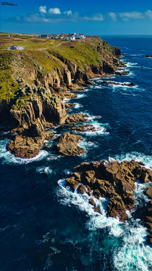 Aerial view of rugged coastal cliffs meeting the ocean, with waves crashing against the rocks. The landscape features green hills and a clear blue sky, showcasing a scenic coastal environment.