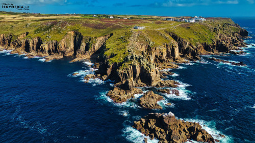 Aerial view of a rugged coastline with cliffs and rocky outcrops, surrounded by deep blue ocean waters. The landscape features green hills and a few white buildings in the distance, under a clear blue sky with scattered clouds.