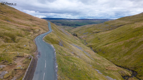 Aerial view of a vast, undulating landscape with green hills, a winding road, and overcast skies.