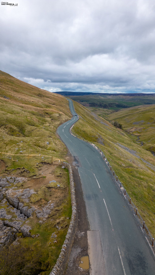 Aerial view of a vast, undulating landscape with green hills, a winding road, and overcast skies.