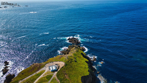 Aerial view of a coastal landscape featuring a rocky cliff extending into the ocean. The vibrant blue water contrasts with the green grass on the cliff. A small building is visible at the cliff's edge, surrounded by scenic views of the sea and distant landforms.