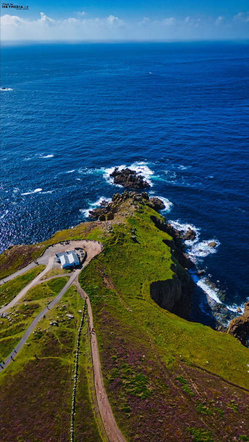 Aerial view of a rocky coastline with a small building on a green hill. The ocean is deep blue, with waves crashing against the rocks. The landscape features a mix of grass and rocky terrain, under a clear blue sky.