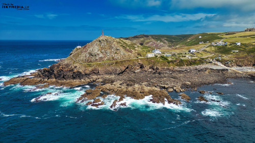 Aerial view of a coastal landscape featuring a rocky shore, rolling green hills, and a lighthouse on a hilltop. The ocean waves crash against the rocks, creating white foam. A few white buildings are scattered along the coastline under a blue sky with some clouds.